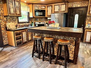 A kitchen with a stone wall, wooden cabinets, a central island with stools, a microwave, a stove, and a refrigerator. - Luxury Treehouse with Soaking Tub near White Mountain National Forest, Stoneham, Maine (Stoneham)