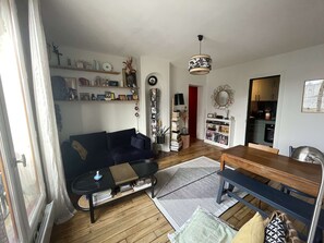 A living room with a sofa, coffee table, and a dining table. There are shelves with books and decorative items. A mirror and a wall clock are also visible. - Parisian apartment on courtyard (Paris)