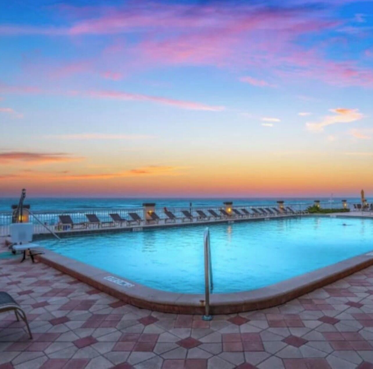 A pool with lounge chairs and a view of the ocean at sunset.