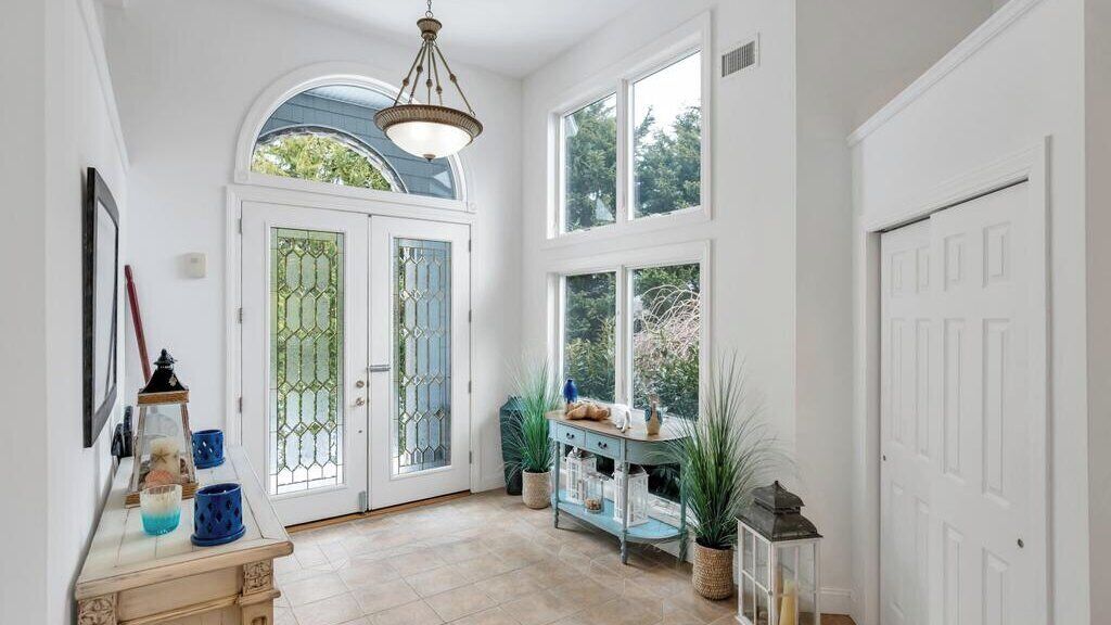 Un pasillo con una puerta blanca, una puerta azul y un ventanal. Hay una mesa con adornos y una planta en maceta.