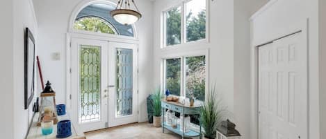 Un pasillo con una puerta blanca, una puerta azul y un ventanal. Hay una mesa con adornos y una planta en maceta.
