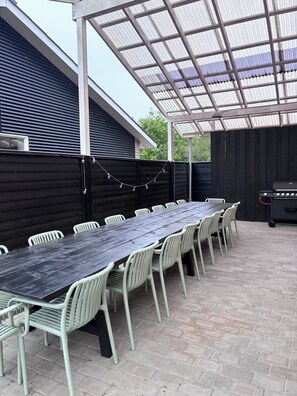 An outdoor patio with a long table and chairs, a grill, and a covered area with a metal roof.