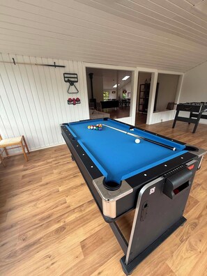 A pool table with balls, a chair, and a billiard cue rack in a room with wooden flooring and white walls.