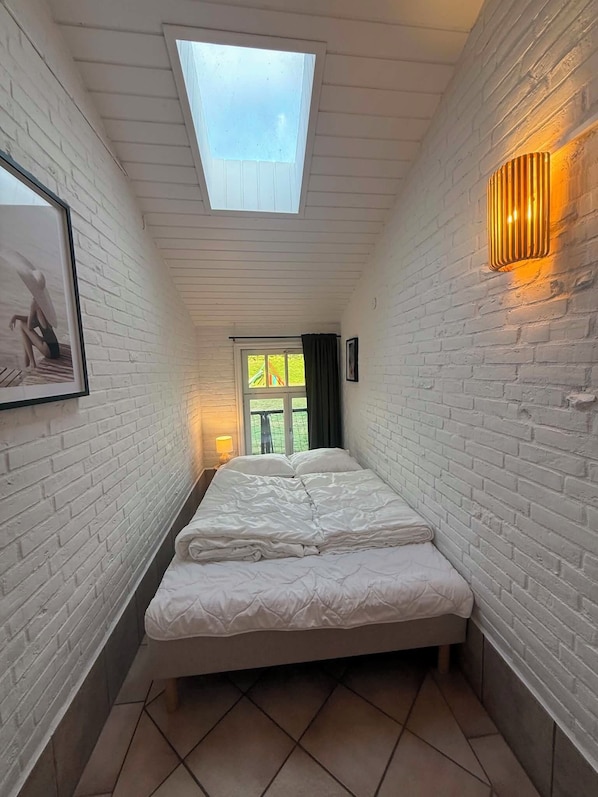 A narrow room with a skylight, a bed, a wall-mounted light, and a framed picture.