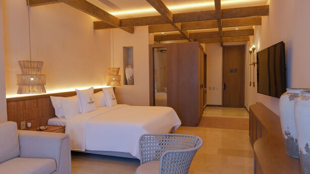 Une chambre d’hôtel moderne dotée d’un grand lit, d’un coin salon avec un fauteuil en osier, d’une armoire en bois et d’un téléviseur à écran plat fixé au mur.
