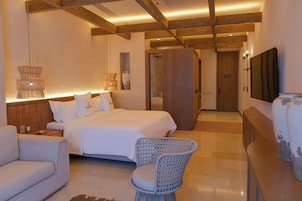 A modern hotel room with a large bed, a seating area with a wicker chair, a wooden wardrobe, and a flat-screen TV mounted on the wall.