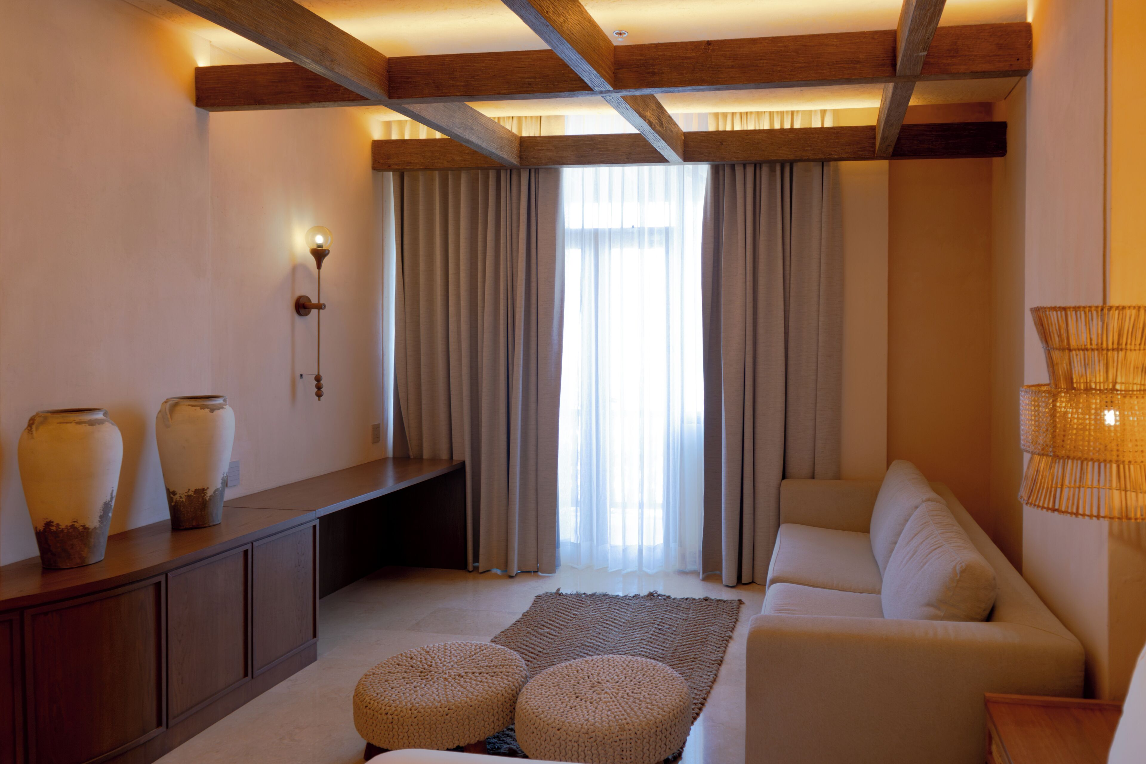 A modern living room with a sofa, ottomans, a console table with vases, and a large window with curtains.