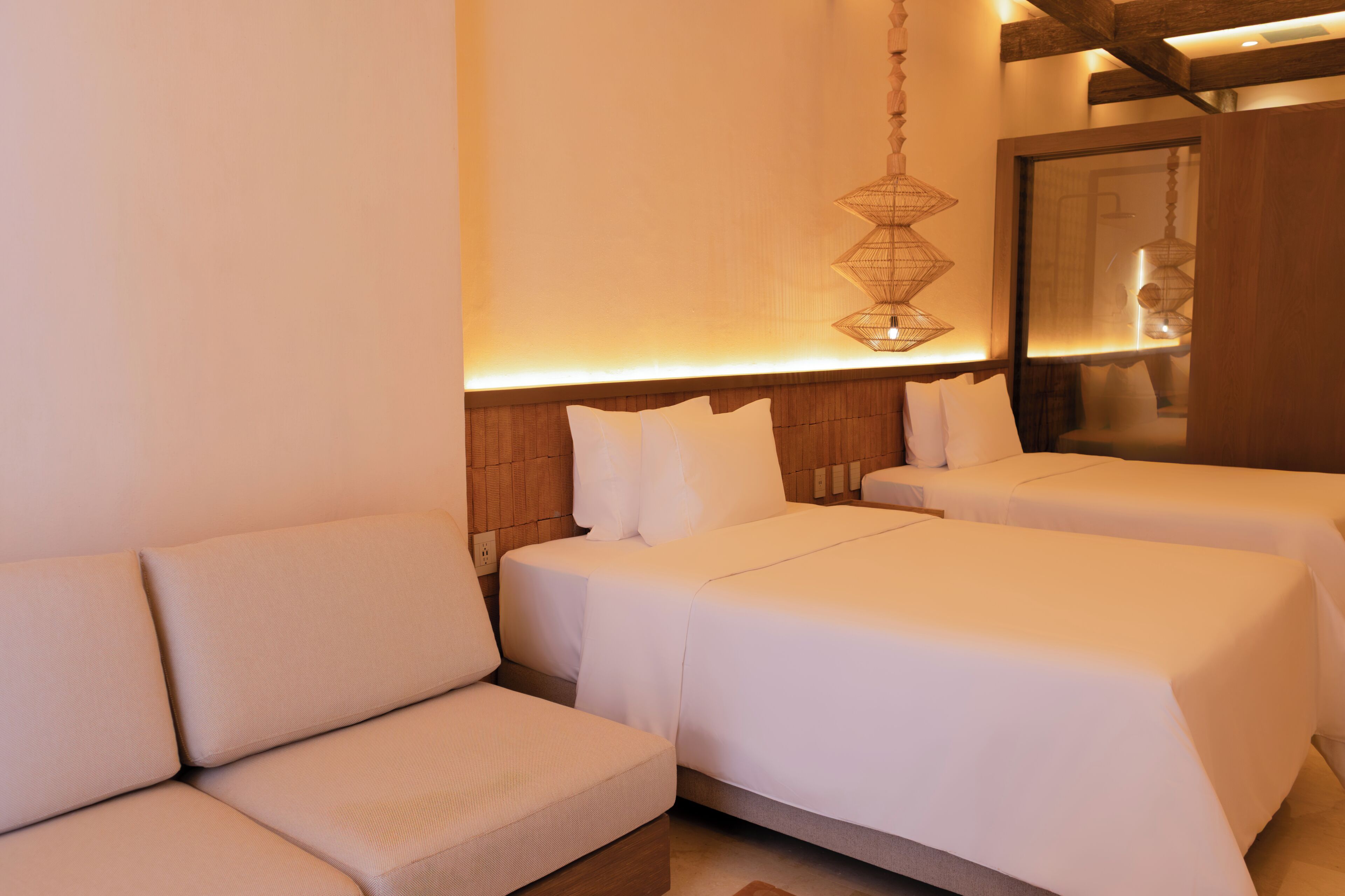 A hotel room with a sofa, two beds, a hanging light fixture, and a wooden headboard.