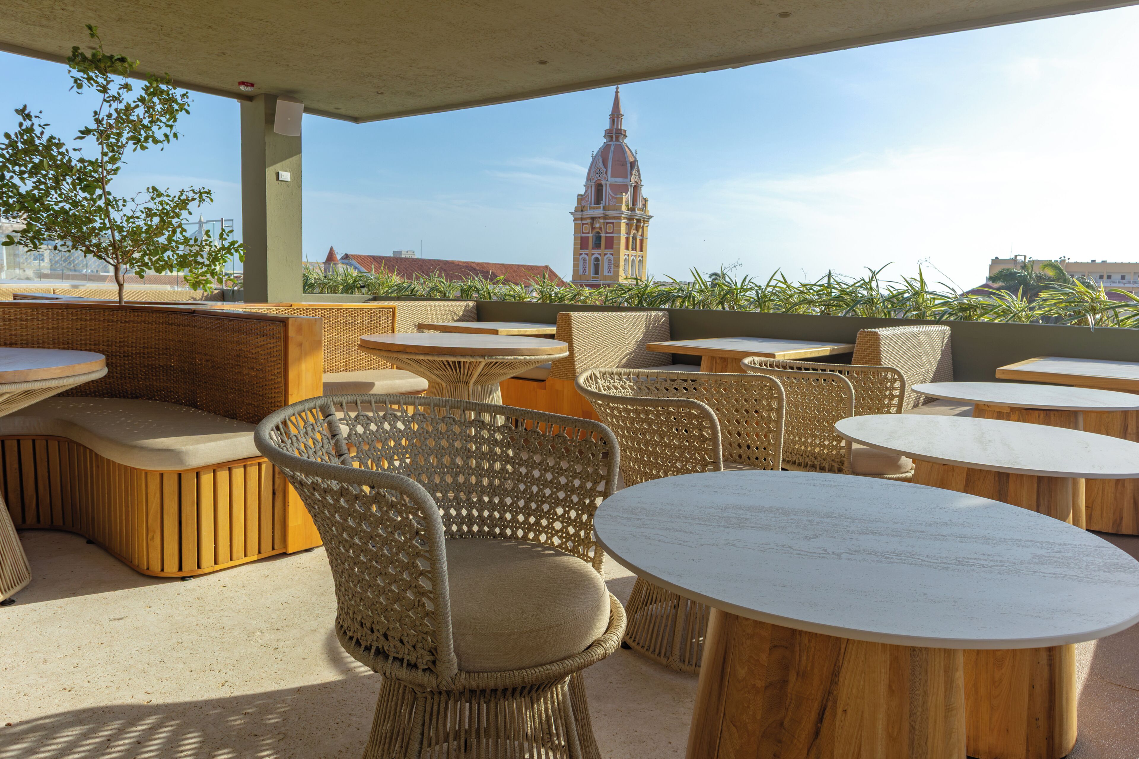 An outdoor seating area with wicker furniture and round tables, overlooking a cityscape with a prominent tower.