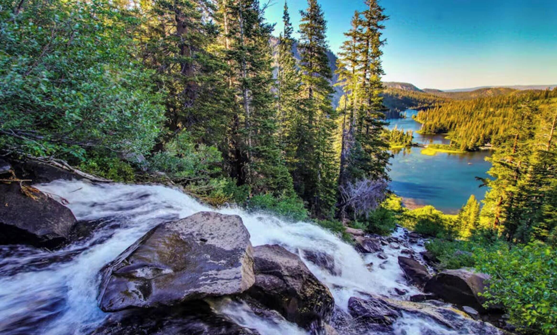 Ein Wasserfall stürzt über Felsen in einem bewaldeten Gebiet mit einem See und Wald im Hintergrund.