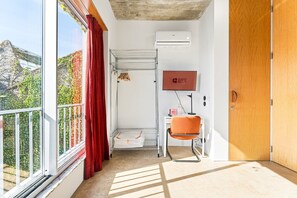 A hotel room with a large window, a red curtain, a white desk with a chair, a mounted TV, and a wooden door. - GuestReady - House of Many Faces - Room 1 (Porto)