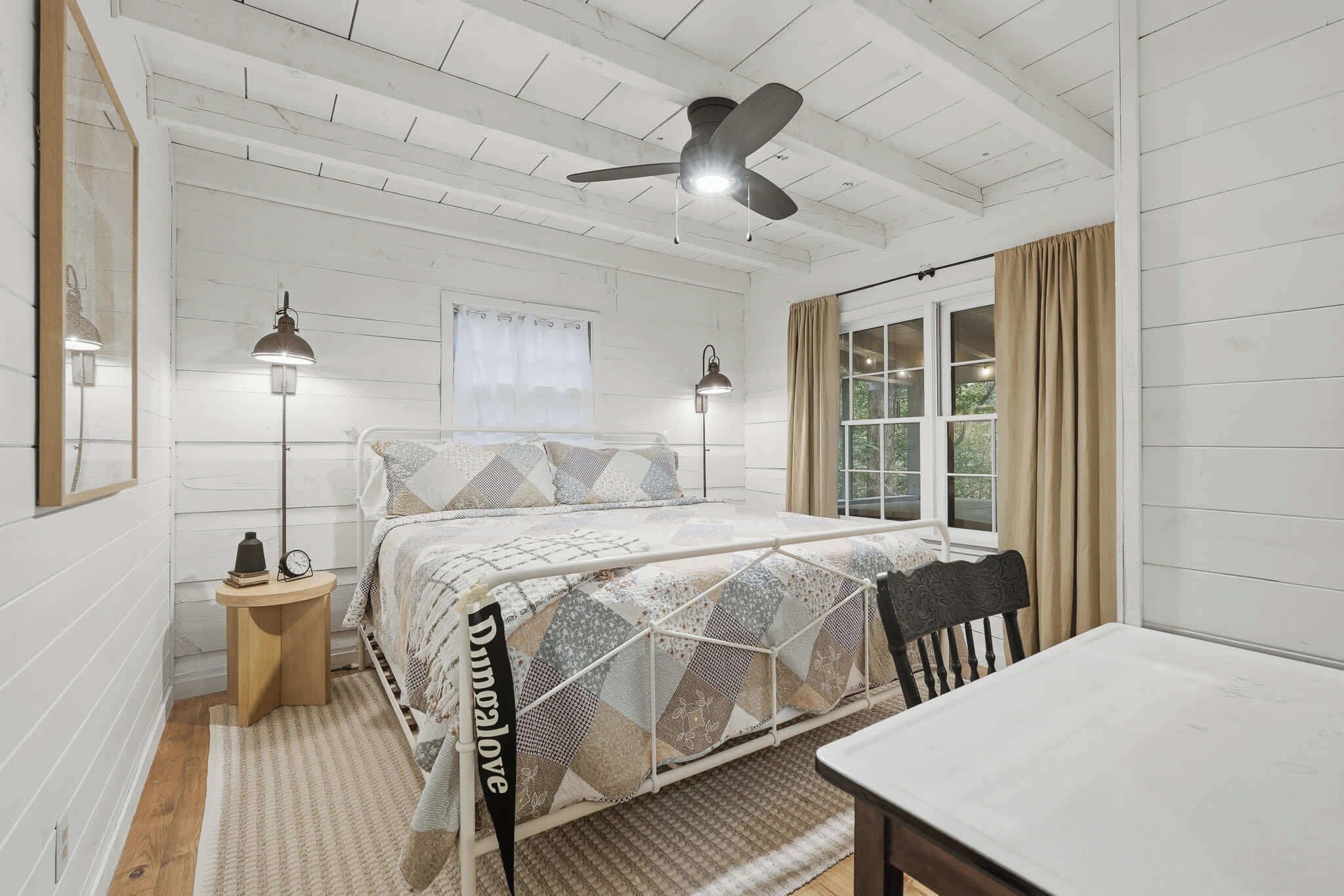 A bedroom with a bed, bedside lamps, a ceiling fan, a window with curtains, and a table.