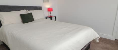 Una cama bien tendida con sábanas blancas, un cabecero oscuro y una mesita de noche con una lámpara roja.