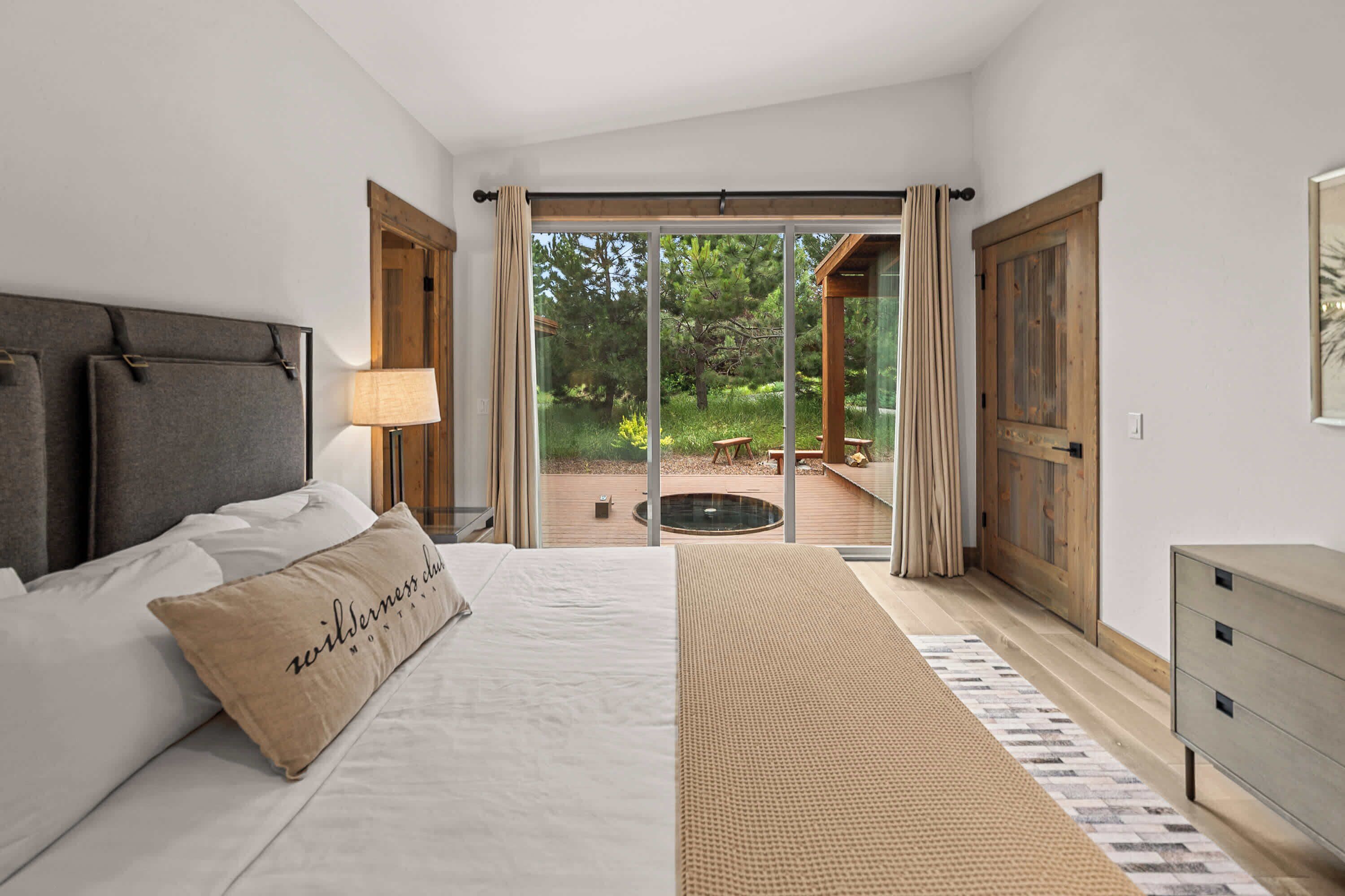 A bedroom with a bed, a nightstand, a lamp, a wooden bench, and a view of an outdoor deck.