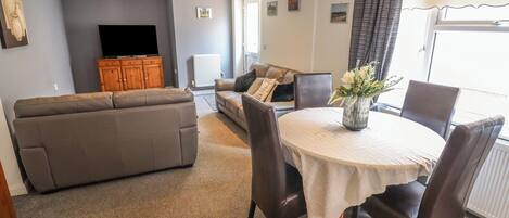 Un salon comprenant un canapé, une table à manger avec des chaises, un téléviseur posé sur un meuble en bois et une fenêtre avec des rideaux.