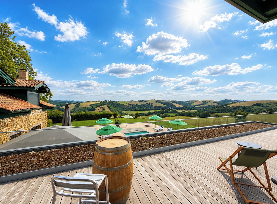 Une terrasse en bois offrant une vue sur des collines verdoyantes, avec des sièges d’extérieur et un tonneau.