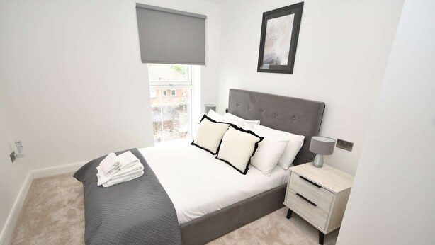 Een slaapkamer met een bed, nachtkastje, lamp, raam met een blind en een schilderij aan de muur.