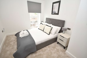 Une chambre à coucher comprenant un lit, une table de chevet, une lampe, une fenêtre avec un store et un tableau au mur.