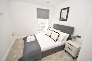 A bedroom with a bed, bedside table, lamp, and a window with a view. - Stylish 1 Bedroom Apartment in Central Leeds (Leeds)