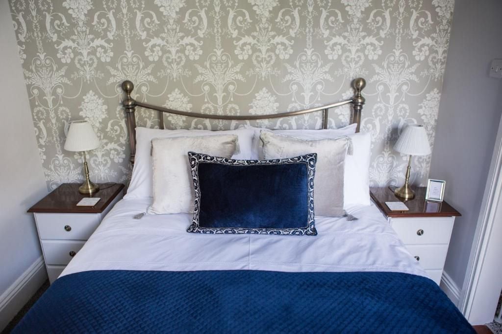 A bed with a white and blue bedspread, two bedside tables with lamps, and a patterned wallpaper background.
