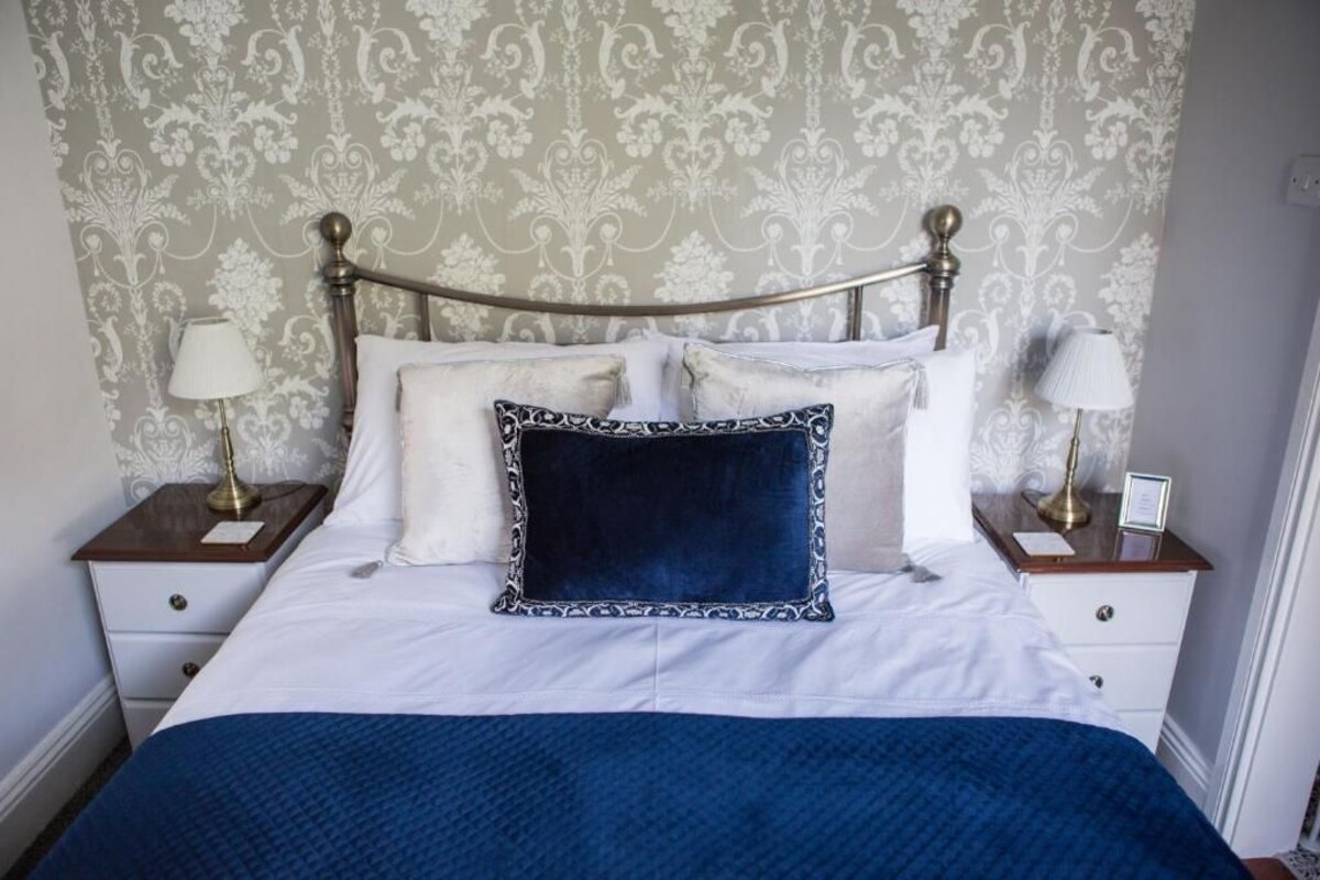 Una cama con una colcha blanca y azul, dos mesitas de noche con lámparas, y un fondo de papel tapiz estampado.