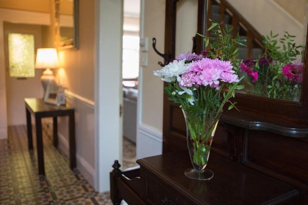 A vase with flowers on a wooden dresser in a room with patterned flooring and a lamp in the background.