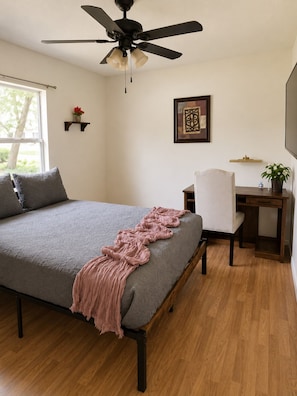 A bedroom with a bed, a desk, a chair, a ceiling fan, a window, and a wall-mounted shelf with a plant.