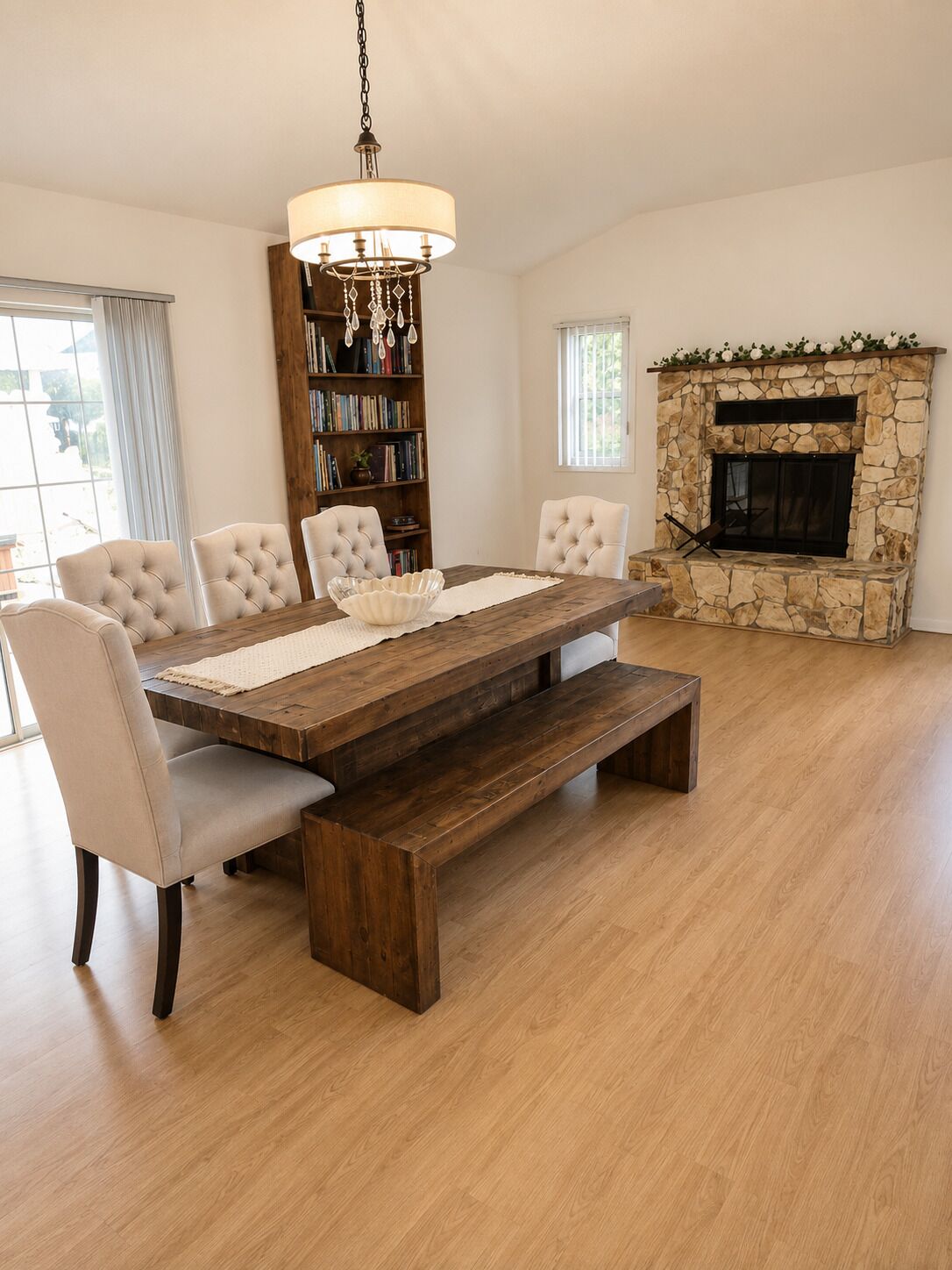 A dining area with a wooden table, cushioned chairs, a chandelier, a stone fireplace, and a bookshelf.