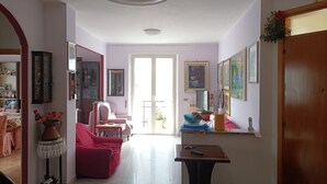 Uma sala de estar ampla com uma grande janela, uma área de jantar com mesa e cadeiras, e um corredor que leva a uma porta.
