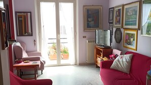 Uma sala de estar com um sofá vermelho, uma poltrona rosa, uma televisão e uma varanda com plantas em vasos.