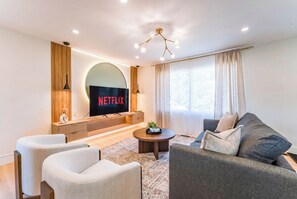 A modern living room with a flat-screen TV displaying 'NETFLIX,' a sofa, two armchairs, a coffee table, and a decorative pendant light. - Luxury Designer Home with Spa Bathrooms • King Bed • AC • Minutes to Downtown (Calgary)