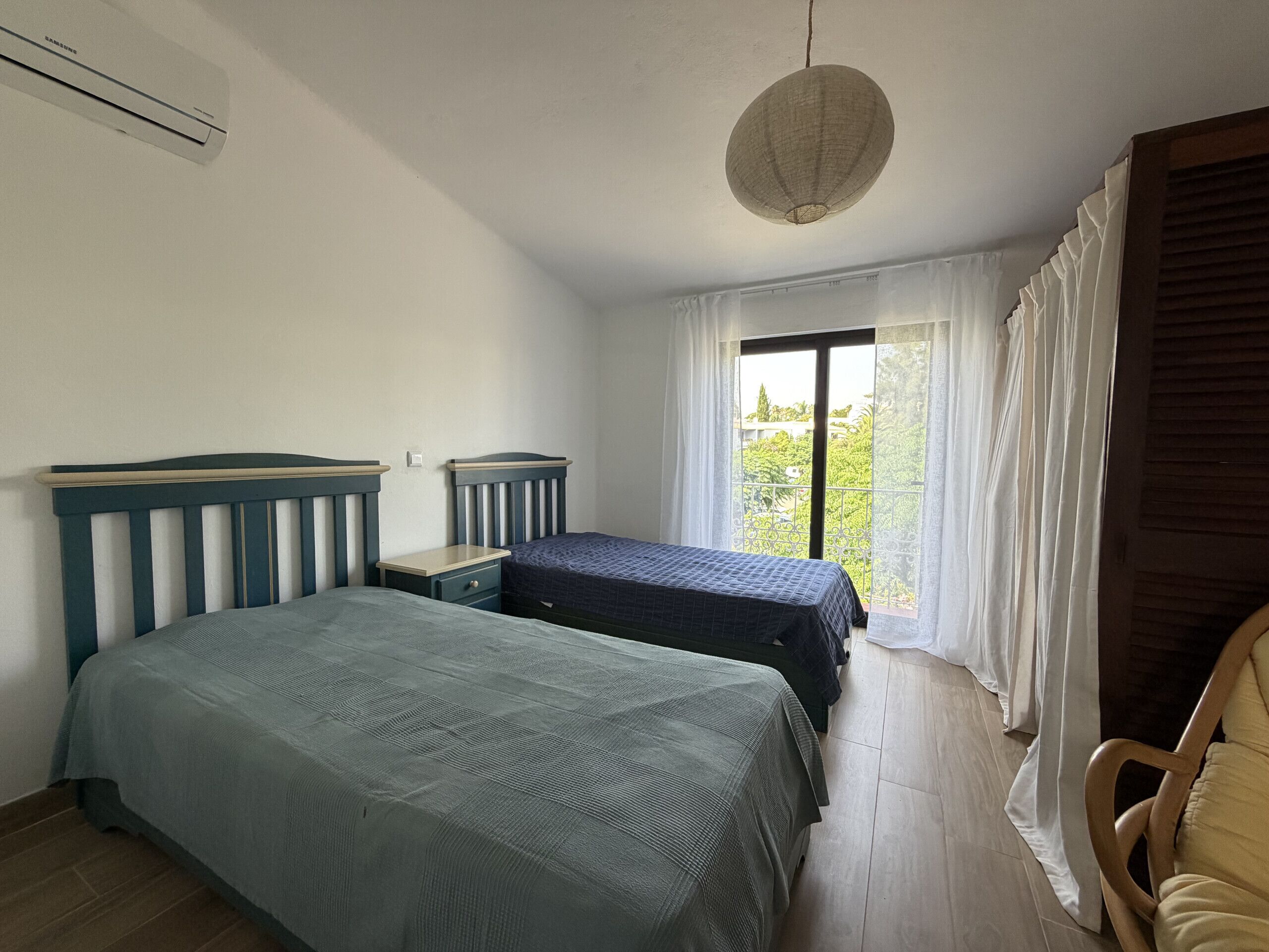 A bedroom with two beds, a window with curtains, a chair, and a hanging light fixture.