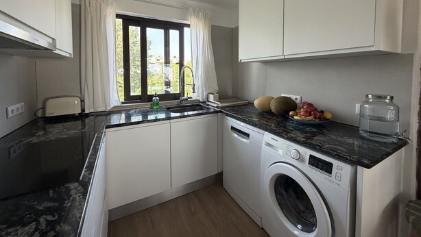 Een compacte keuken met een ingebouwde wasmachine, een raam met gordijnen en een aanrecht met fruit en een waterfles.