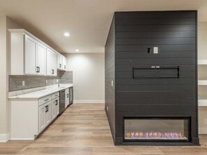 A modern kitchen with white cabinetry, a grey backsplash, and a black fireplace with a visible fire.