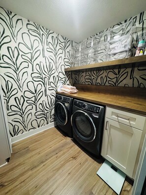 A laundry area with a washing machine, dryer, and a shelf above containing folded towels and toiletries.