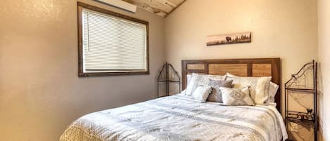 Une chambre avec un plafond en bois, un lit avec une literie à motifs, une fenêtre avec des stores et une étagère fixée au mur.