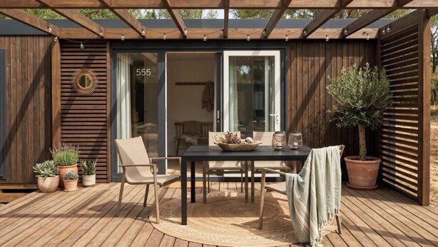A wooden deck with a table and chairs, potted plants, and a sliding glass door leading to a room.