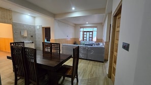 A spacious living area with a wooden dining table and chairs, a kitchen area with a built-in stove, and a large window providing natural light. - Rajguru Guest House (Ujjain)