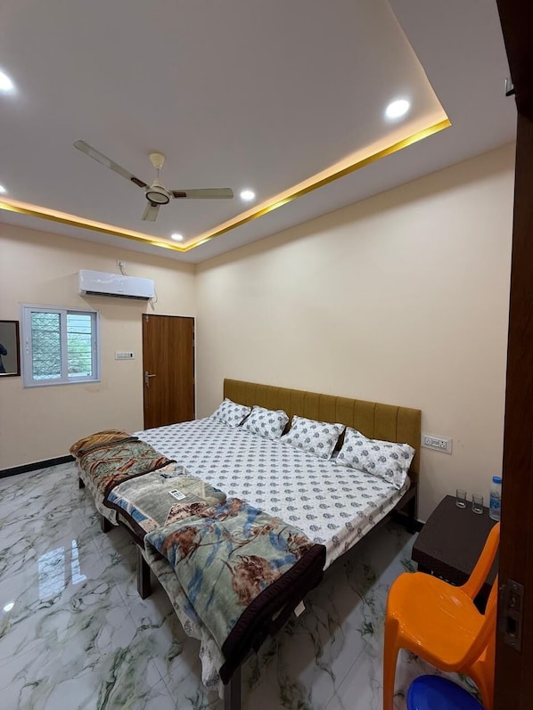 A bedroom with a bed, a ceiling fan, an air conditioning unit, a wooden wardrobe, and a small table with a chair. - Rajguru Guest House (Ujjain)