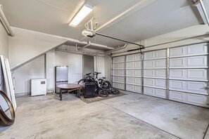 Uma garagem com piso de concreto, ventilador no teto, geladeira, mesa e duas bicicletas.