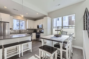 A modern kitchen with white cabinetry, stainless steel appliances, and a dining area with white chairs and a dark wooden table. - Walk to Olde Town Arvada | 2 Ensuite Bedrooms | Garage | Sleeps 5 (Arvada)