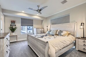 Une chambre à coucher comprenant un lit, un ventilateur de plafond, une table de chevet, une lampe, une commode, une fenêtre avec des stores et une plante.