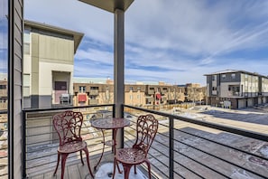 Uma varanda com duas cadeiras de metal decoradas e uma pequena mesa redonda, com vista para uma área residencial com edifícios altos.