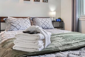 A neatly made bed with a stack of folded towels, a green blanket, and a nightstand with a clock displaying 10:56. - 3BR Westminster Home with Hot Tub, Yard + King Suite (Westminster)