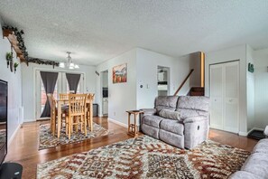 A living room with a grey sofa, wooden dining table, and chairs. There is a staircase leading to another level, and a white door with a green sign. - 3BR Westminster Home with Hot Tub, Yard + King Suite (Westminster)