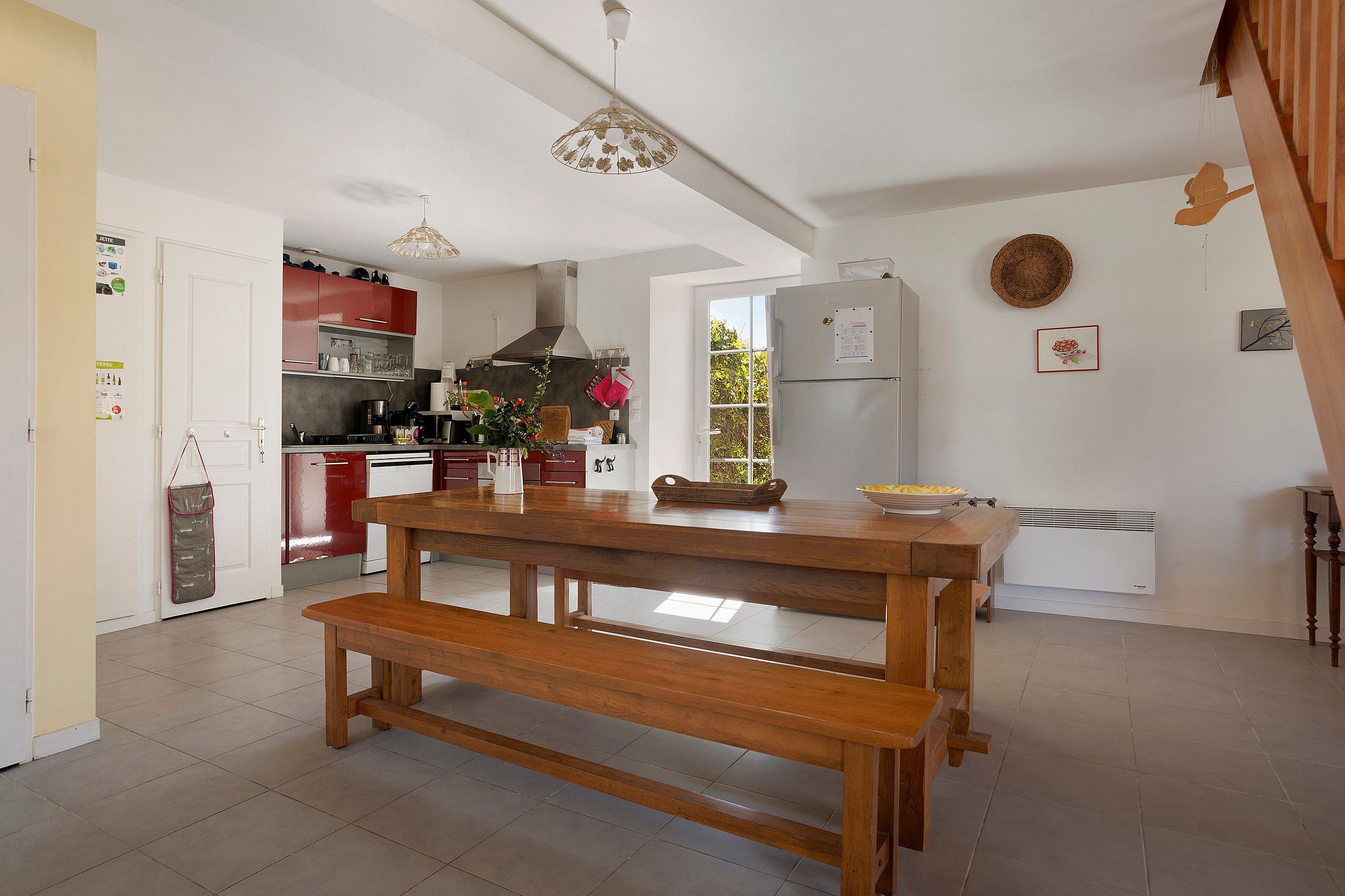Een keuken met een houten tafel en bank, een koelkast en een raam met uitzicht op groen.