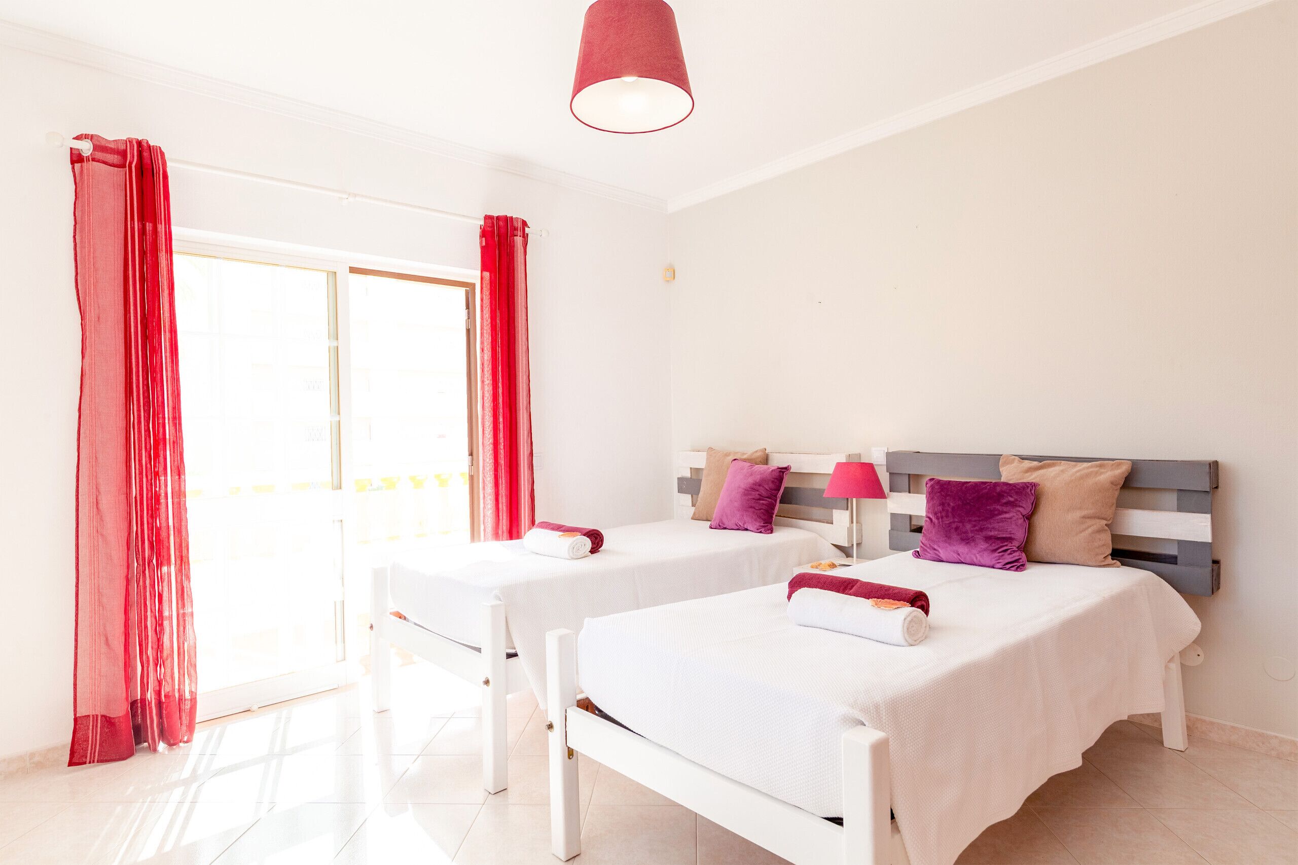 A hotel room with two single beds, a red curtain, a white ceiling lamp, and a view of the outside.