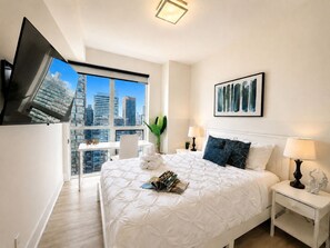 A modern hotel room with a large bed, a TV mounted on the wall, a city view, and a small table with a plant.