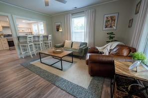 A modern living room with a brown leather sofa, a wooden coffee table, and a green armchair. There is a kitchen area visible in the background.