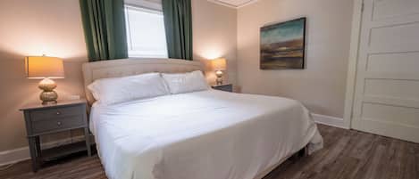 A neatly made bed with white linens, two bedside lamps, a nightstand, a window with green curtains, and a painting on the wall.
